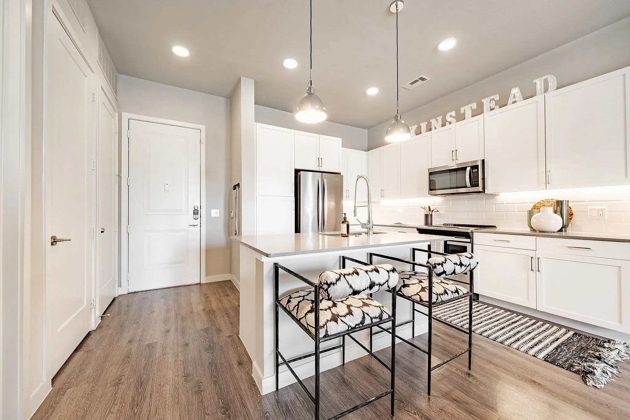 A modern kitchen with white cabinets, a stainless steel refrigerator, a textured backsplash, and stylish bar stools at the countertop.