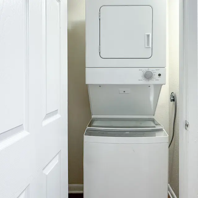 A compact laundry area featuring a stacked washer and dryer unit positioned against a light-colored wall. A closed white door is visible to the left.