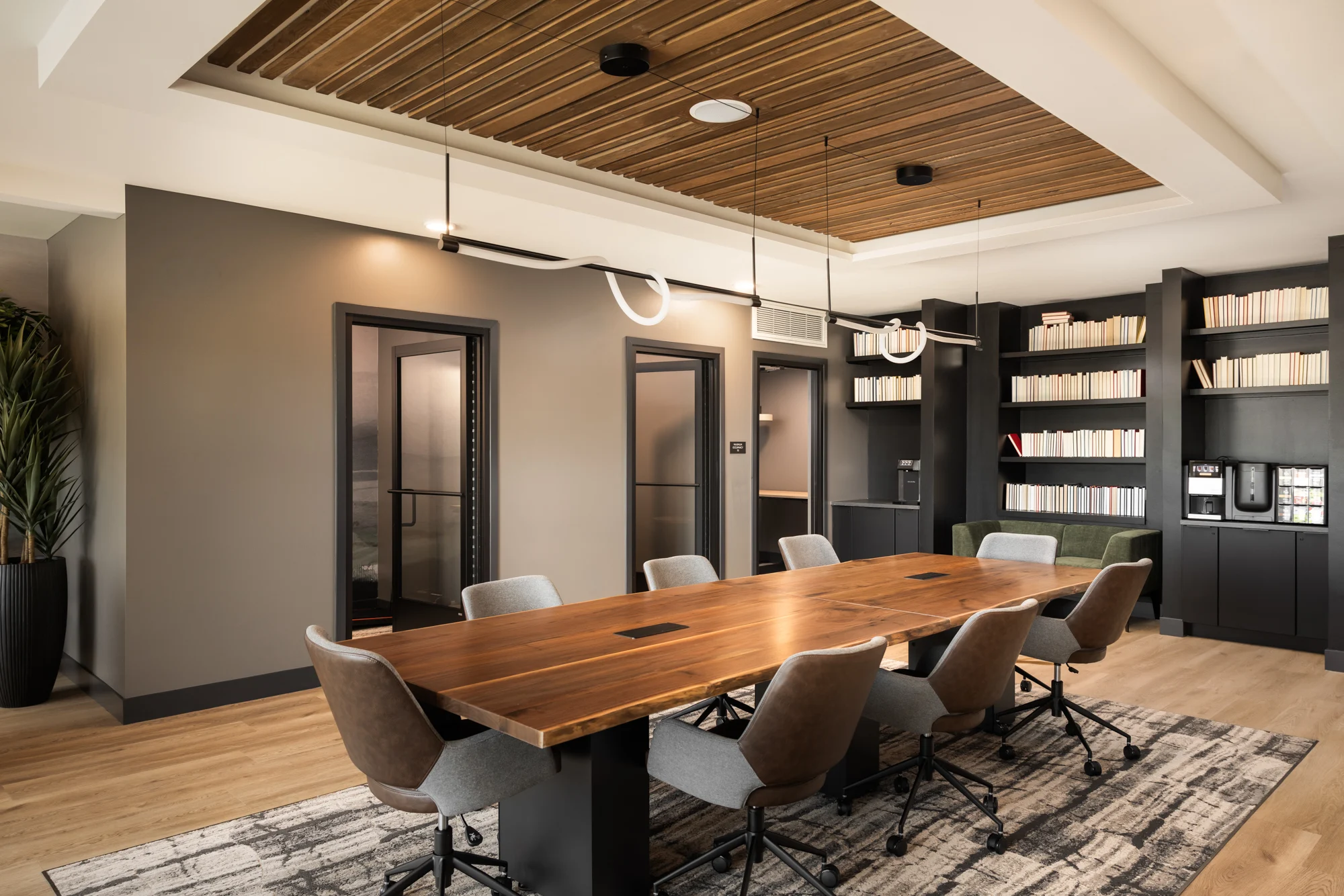 Stylish Conference Room A modern conference room featuring a large wooden table surrounded by ergonomic chairs, wooden paneling on the ceiling, and shelves filled with books.