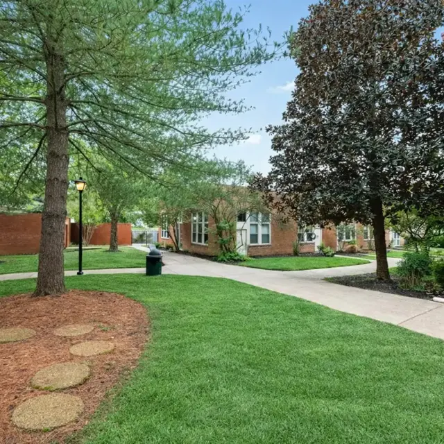 A serene outdoor pathway surrounded by grass, trees, and buildings, featuring stepping stones and lampposts.