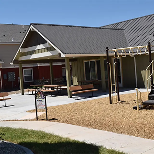 A playground with a covered seating area and benches, surrounded by gravel and grass. In the background, there are residential buildings with different colored facades.