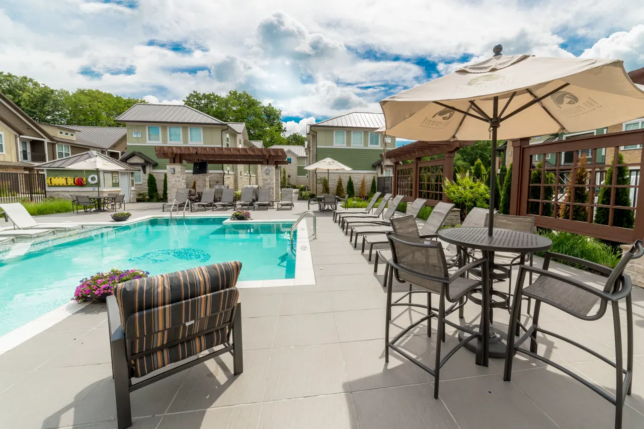 A well-lit swimming pool area with lounge chairs and umbrellas, surrounded by greenery and residential buildings.
