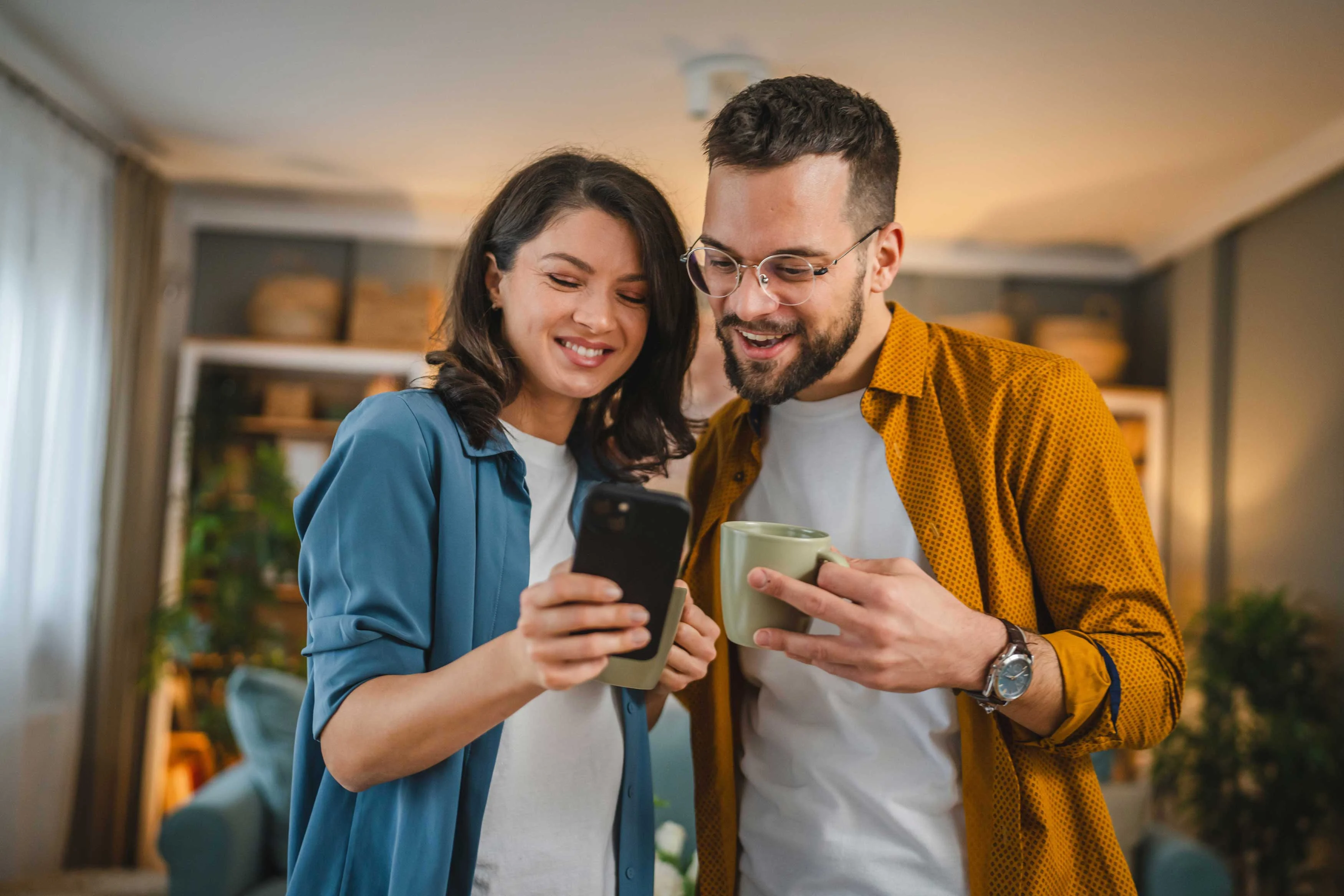 A happy couple looking at a smartphone together, holding cups in a cozy indoor setting.