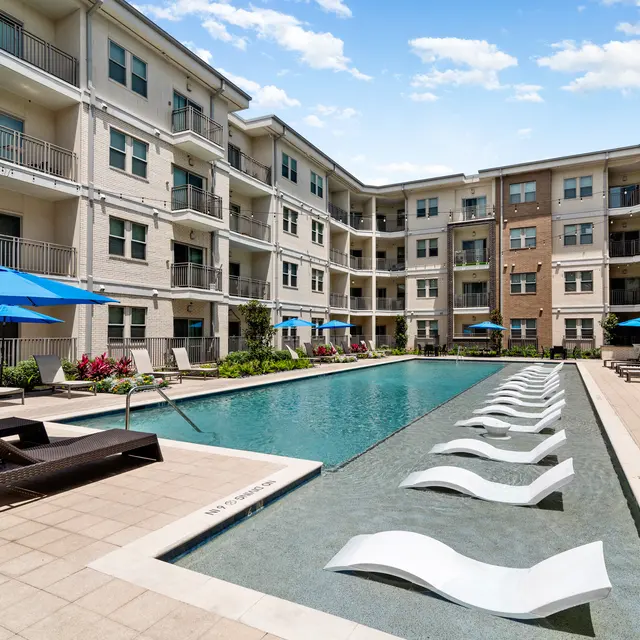A modern apartment complex with a large outdoor swimming pool surrounded by lounge chairs and umbrella shades. The pool is flanked by green landscaping and several buildings.