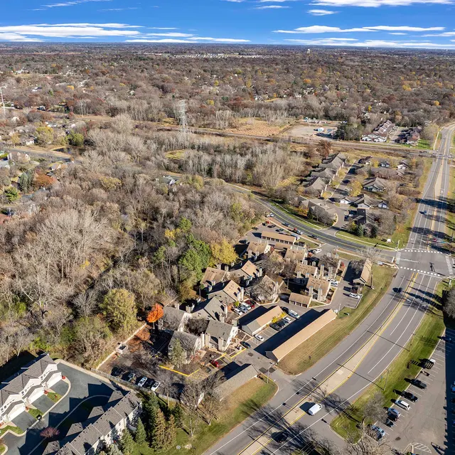 Aerial view of a suburban area featuring residential homes, trees, and streets. The landscape includes a mix of housing developments and open fields, showcasing a blend of wooded areas and organized neighborhoods.