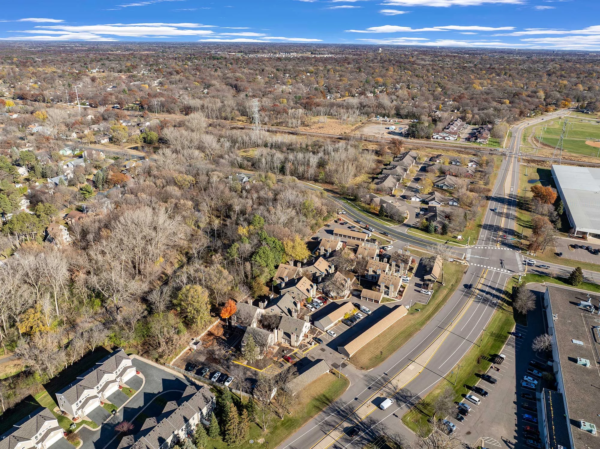 Aerial View of Suburban Neighborhood Aerial view of a suburban area featuring residential homes, trees, and streets. The landscape includes a mix of housing developments and open fields, showcasing a blend of wooded areas and organized neighborhoods.