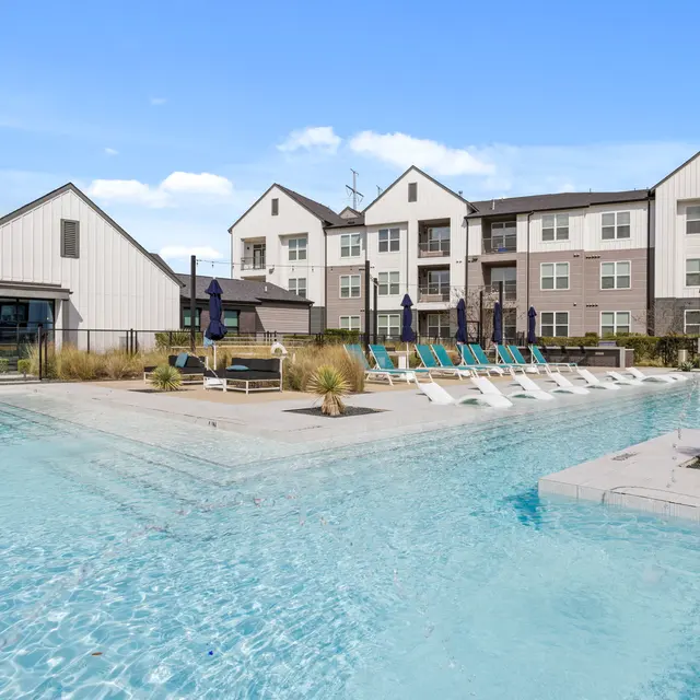 A modern swimming pool area surrounded by lounge chairs and palm plants, with contemporary apartment buildings in the background under a clear blue sky.