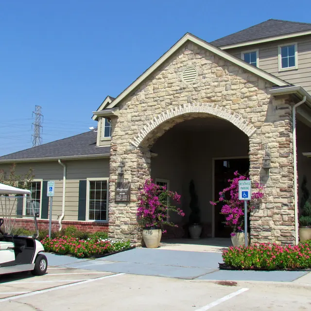 A large building with a stone facade, featuring an entrance archway. A golf cart is parked in front on a paved surface, surrounded by greenery and flowers. Power lines are visible in the background under a clear blue sky.