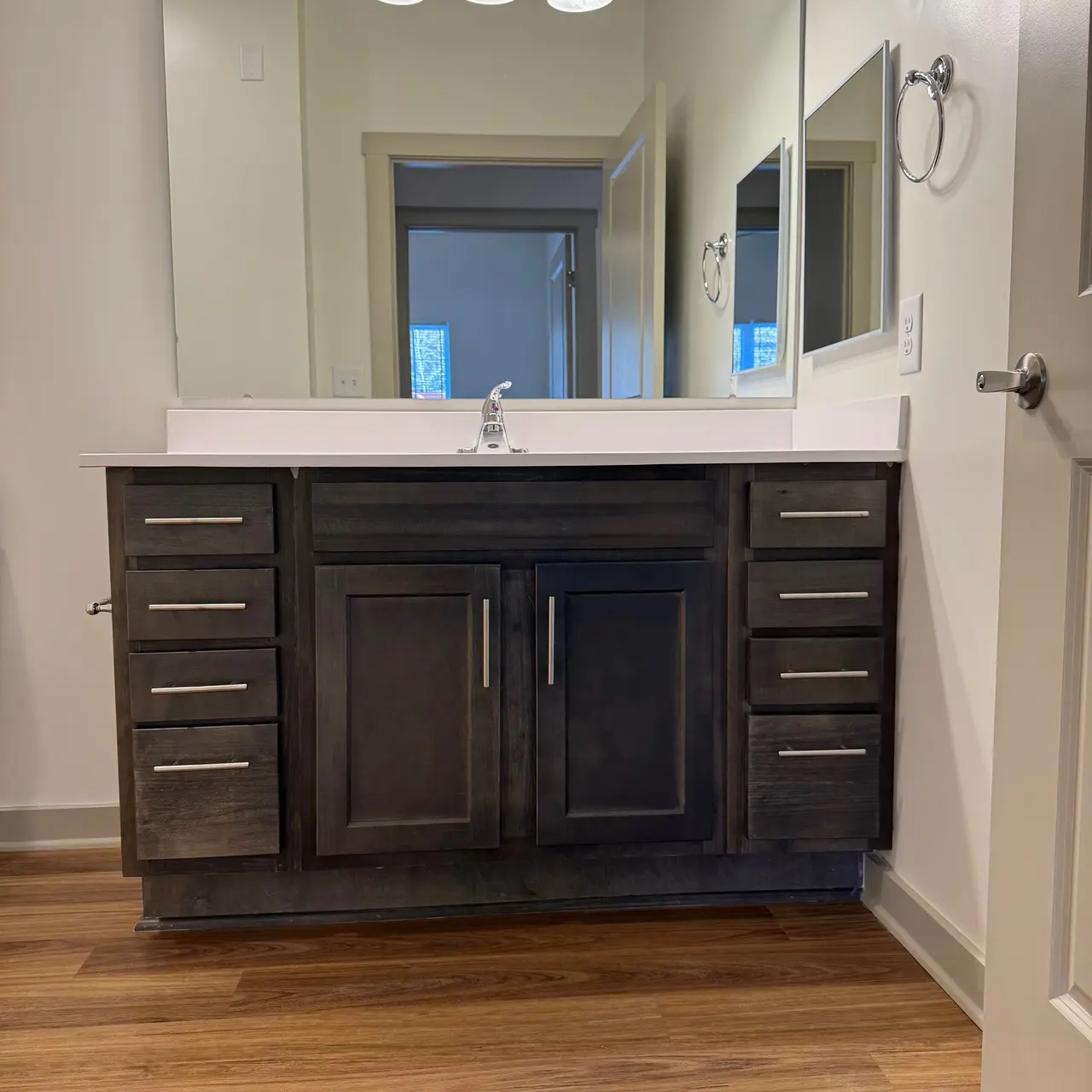 A modern bathroom vanity with a dark wood finish, featuring six drawers and two cabinet doors. A white countertop holds a single faucet, and three light fixtures hang above a large mirror. In the background, a doorway leads to another room with light coming through a window.