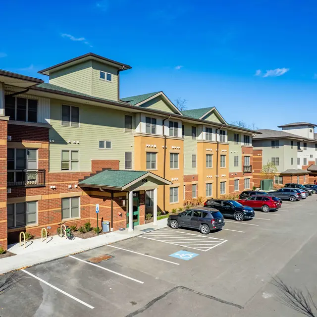 Modern Apartment Complex A modern apartment complex featuring brick and siding exterior, surrounded by a parking lot with several cars parked. Clear blue sky overhead.