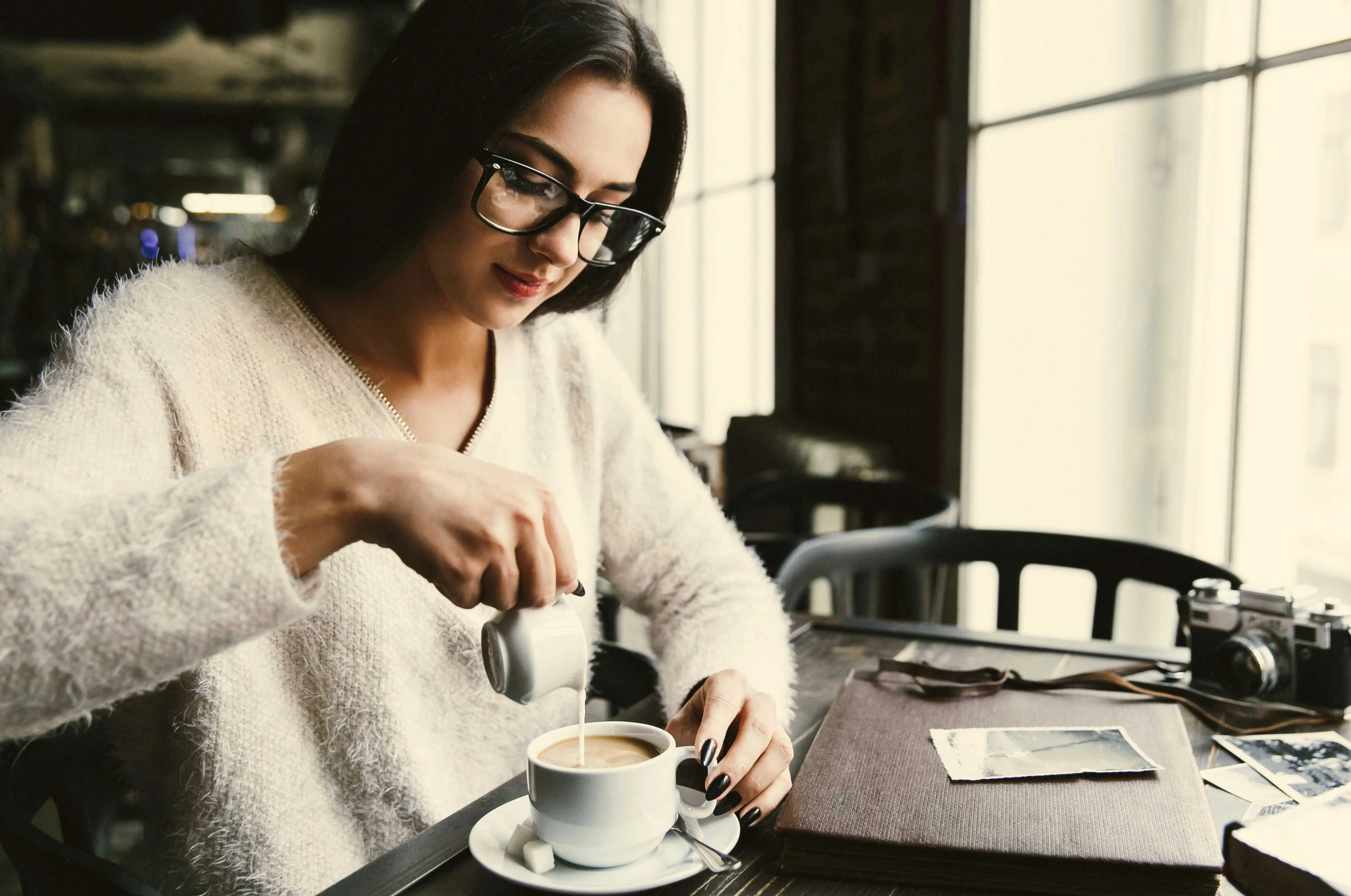 Coffee Moment at Café A woman in a cozy sweater sitting at a café table, pouring cream into a cup of coffee. The setting includes large windows and a camera near her, with some photographs on the table.