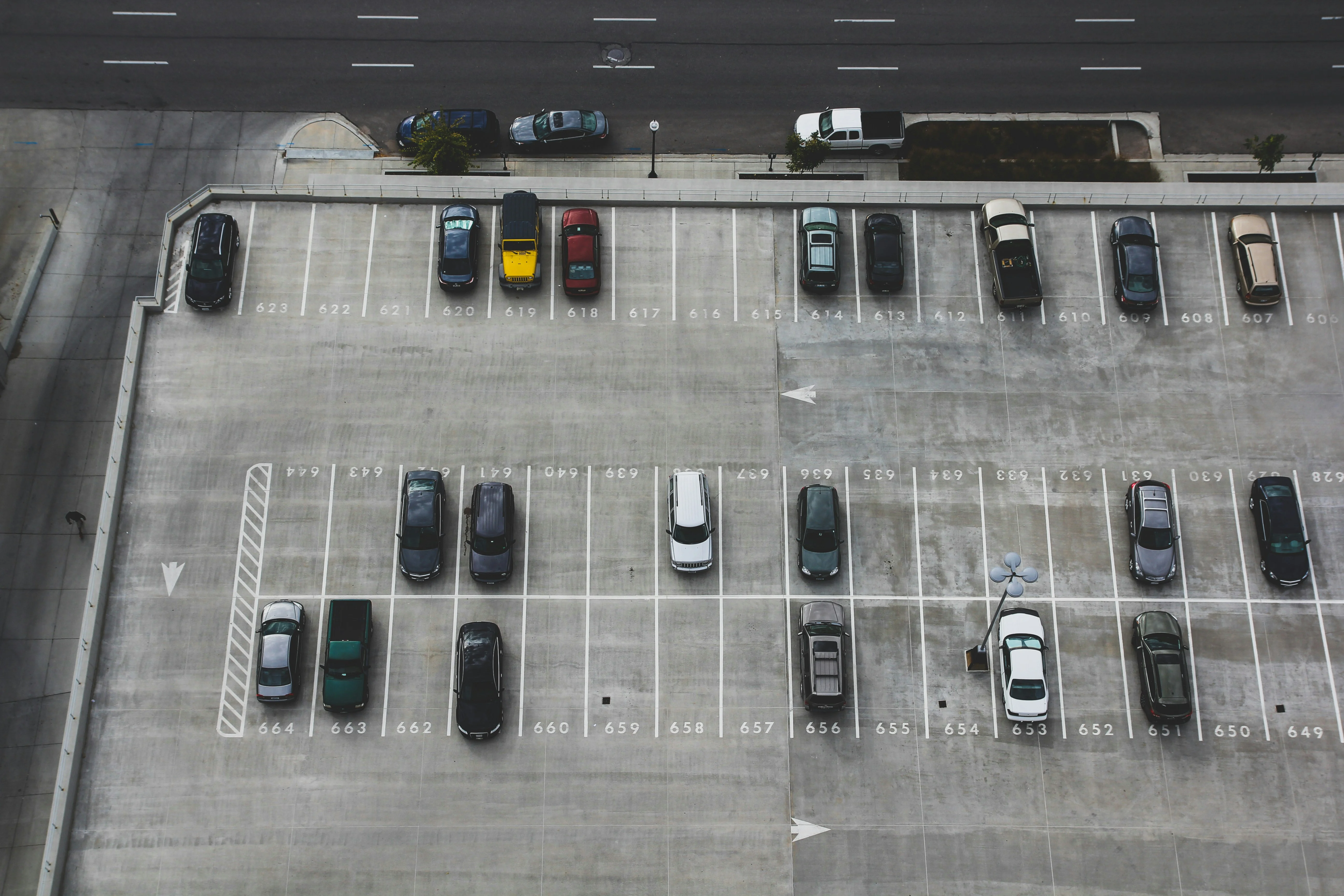 Aerial view of a nearly empty parking lot with various cars parked in designated spaces. A street is visible in the background.