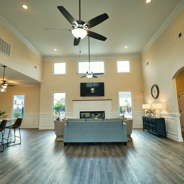 A spacious and modern living room featuring a light-colored sofa, a fireplace, and large windows that provide abundant natural light. The room includes a ceiling fan, a wall-mounted TV, and a dining area with bar stools in the background.
