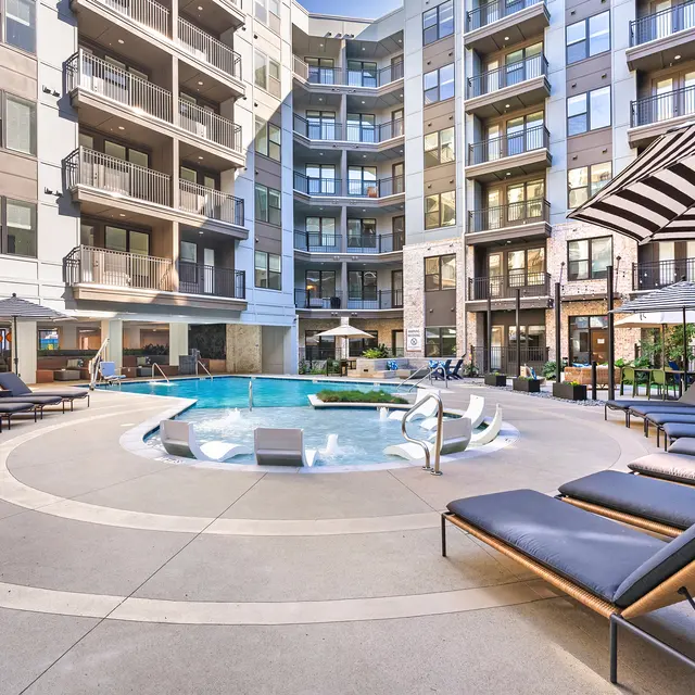 A modern apartment complex featuring a circular swimming pool surrounded by lounge chairs and umbrellas.
