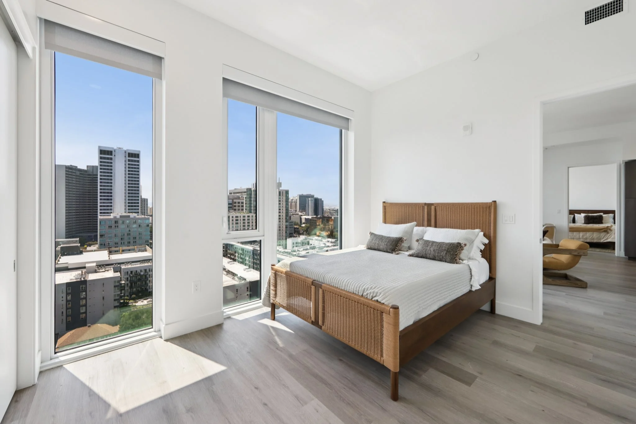 A modern bedroom featuring a bed with decorative pillows, large windows showing a city skyline, and light wood flooring.