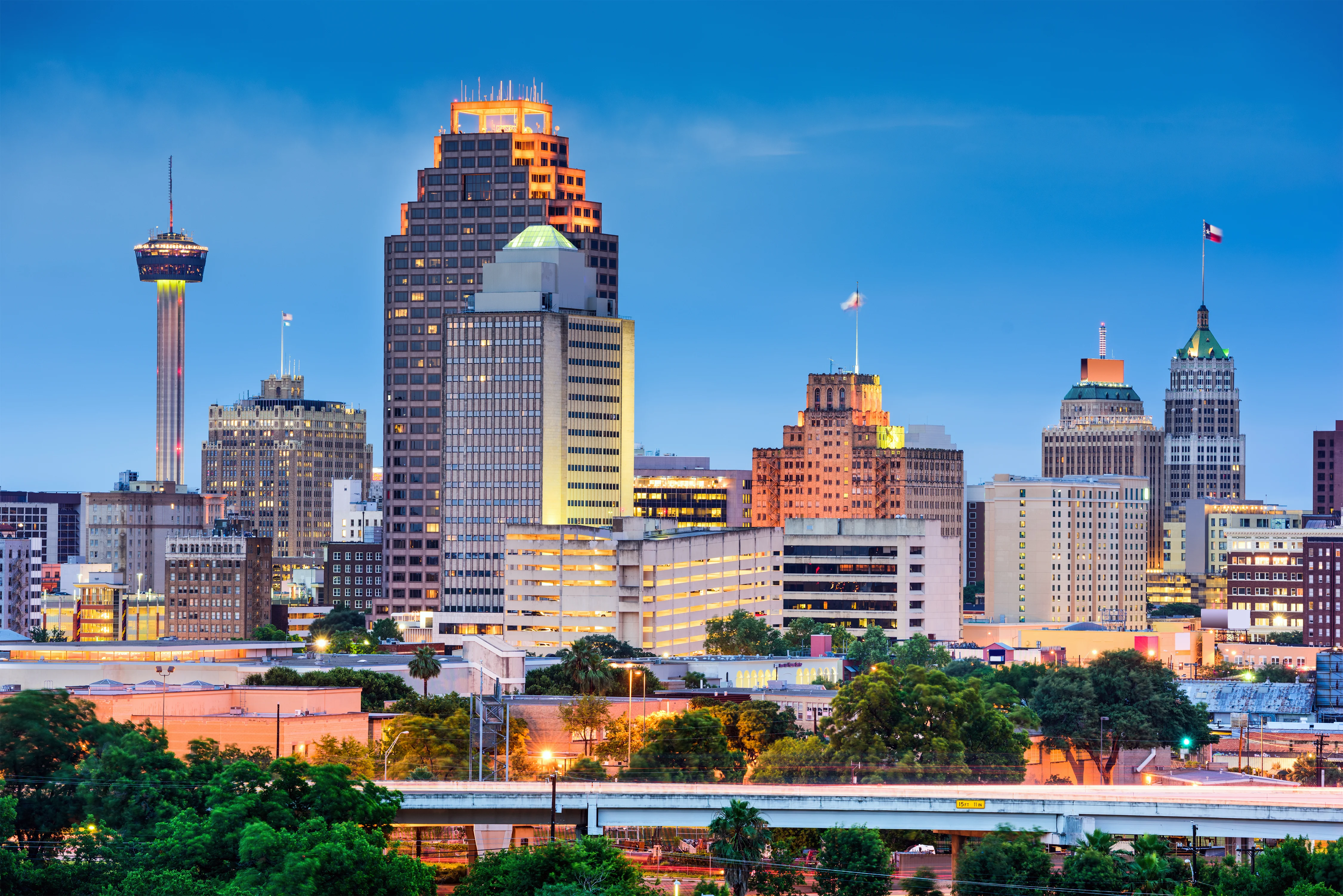 A vibrant skyline view of San Antonio, Texas at dusk, featuring notable buildings illuminated in various colors, with the Tower Life Building and the San Antonio Tower in the background.