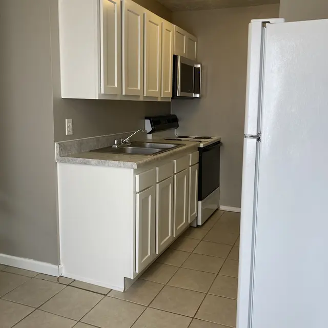 A modern kitchen featuring white cabinets, a gray countertop, a stove, and a stainless steel microwave. The floor is tiled and a white refrigerator is visible at the side.
