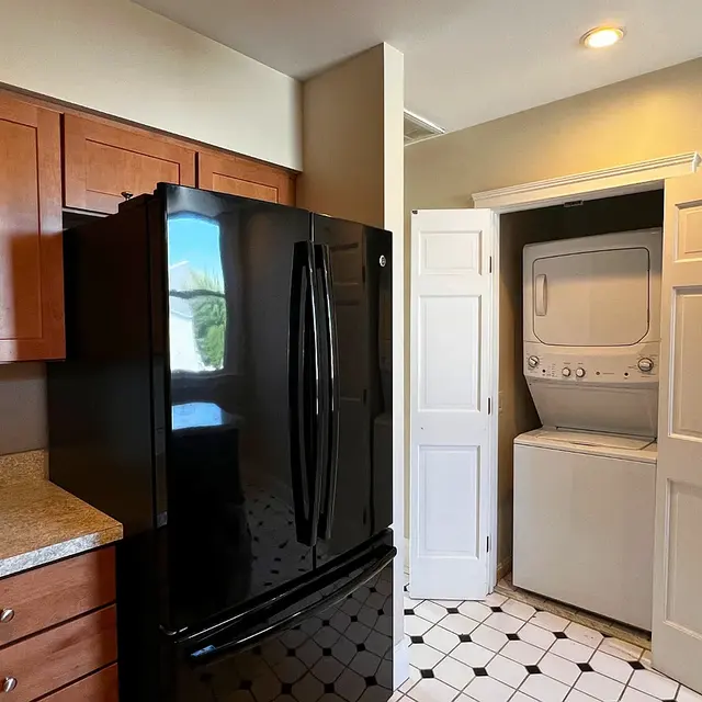 A modern kitchen featuring wooden cabinets, a black refrigerator, and a laundry area with a stacked washer and dryer behind a door.