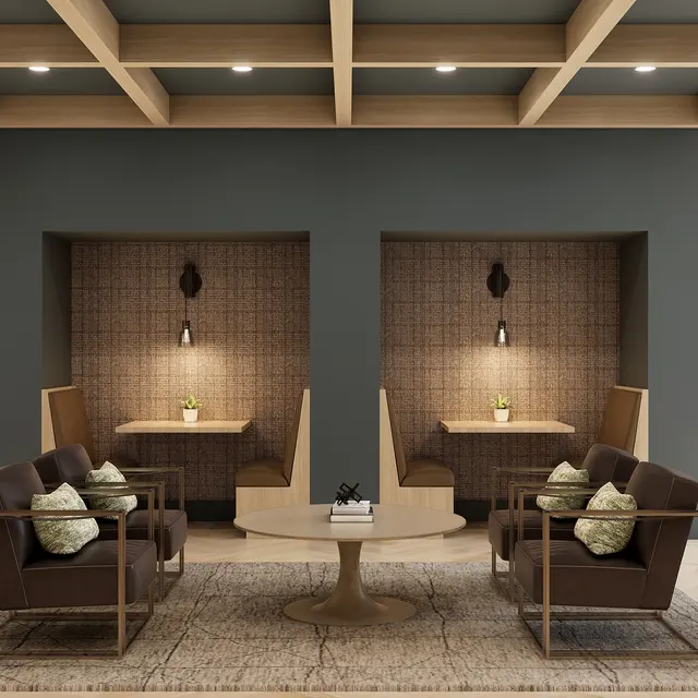 A modern lobby area featuring two cozy seating nooks with brown chairs and a round table. The walls are painted gray, and the ceiling has wooden beams. Soft lighting and decorative plants are present in the background.