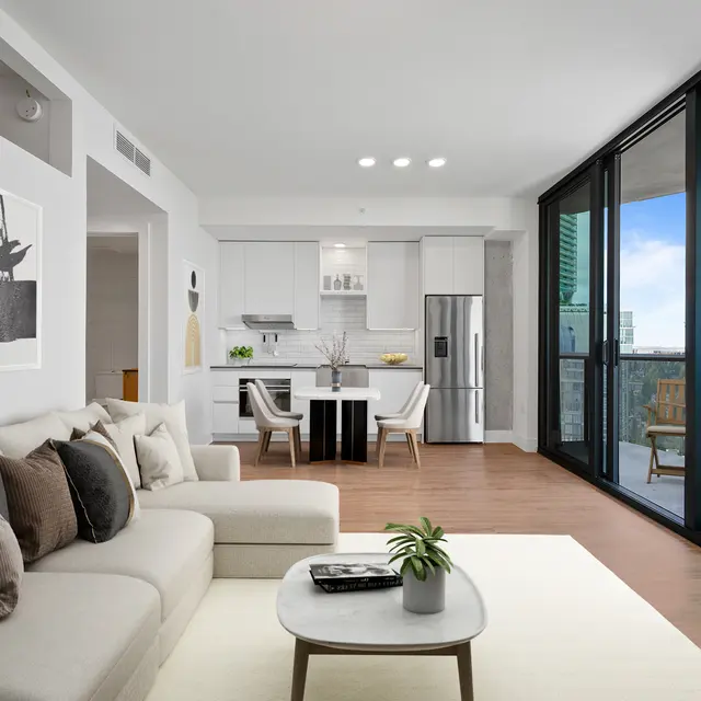 A modern living room featuring a light-colored sofa, a coffee table, and decorative pillows. The space has large glass doors leading to a balcony, and a minimalistic kitchen visible in the background.