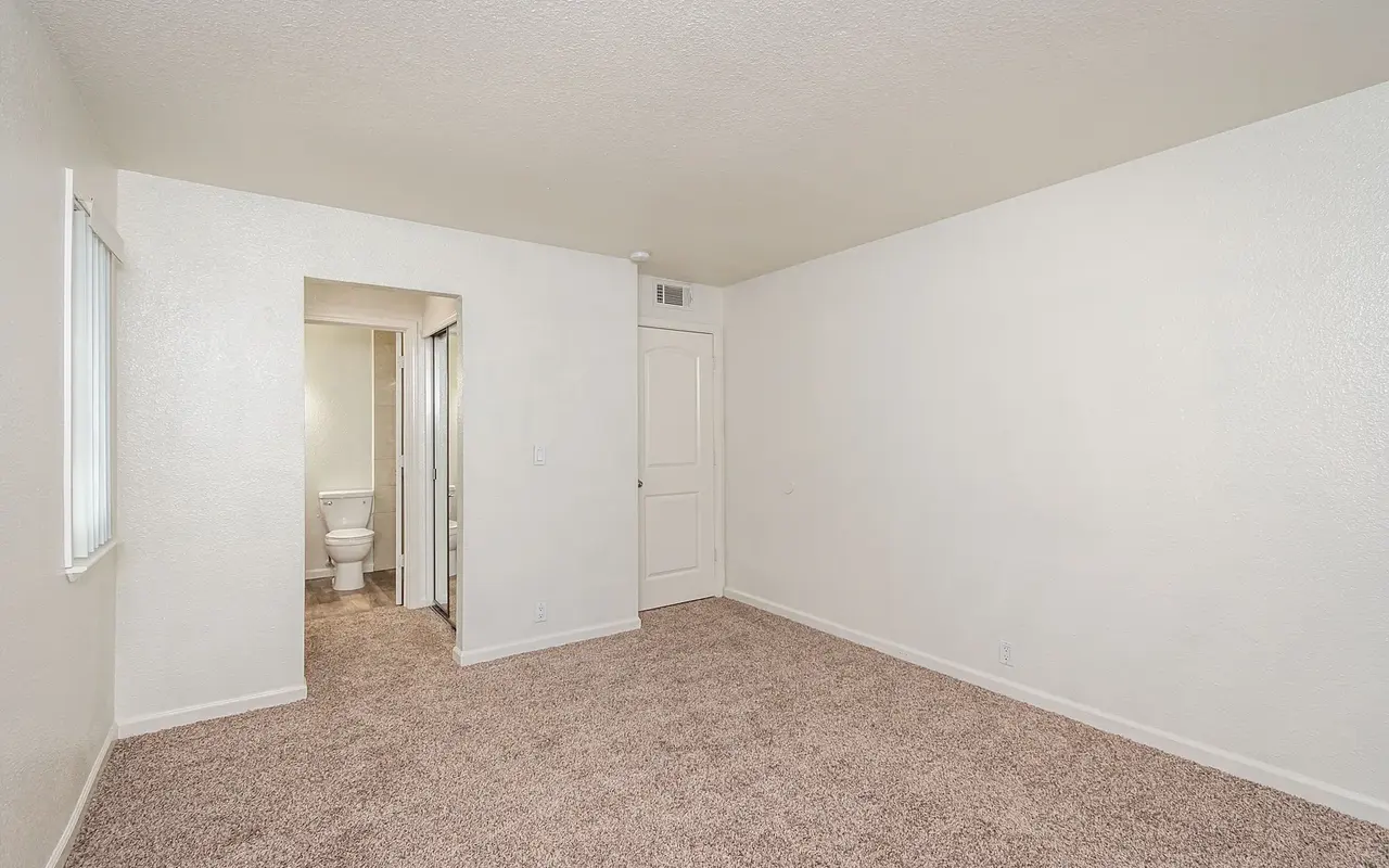 A spacious, empty room with light-colored walls and carpet, featuring a doorway leading to a bathroom.