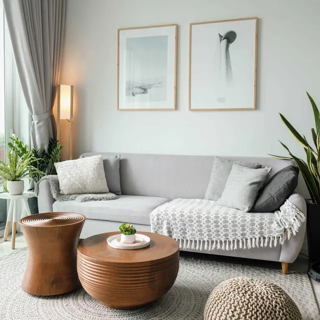 A modern living room featuring a gray sofa with a knitted blanket, a round wooden coffee table with a small plant on it, a knitted pouf, and decorative wall art, alongside indoor plants and curtains.
