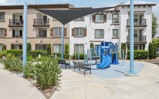 A playground area featuring a blue slide and climbing structure, surrounded by benches and greenery, with a shade sail overhead.