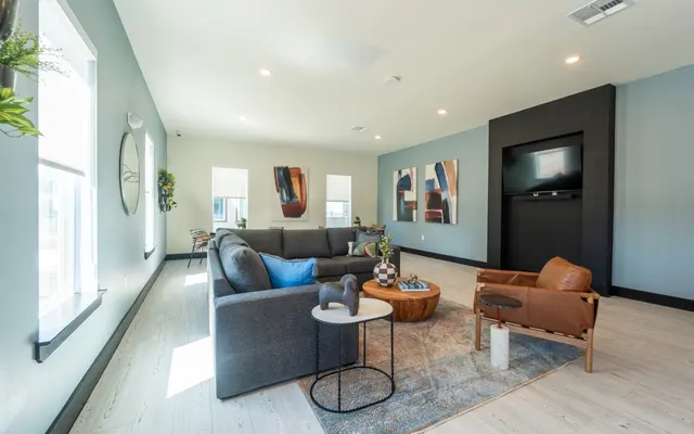 A modern living room featuring a gray sectional sofa with a blue accent pillow, a round coffee table adorned with decorative items, and an orange leather armchair. The walls are painted in light blue, and there are abstract art pieces on the walls. Large windows let in plenty of natural light.