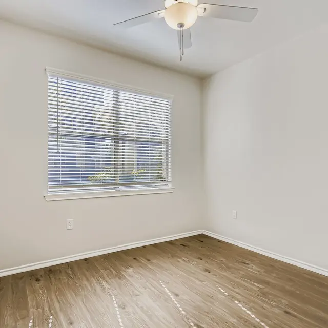 An empty room featuring wooden flooring, a ceiling fan, and a window with blinds.