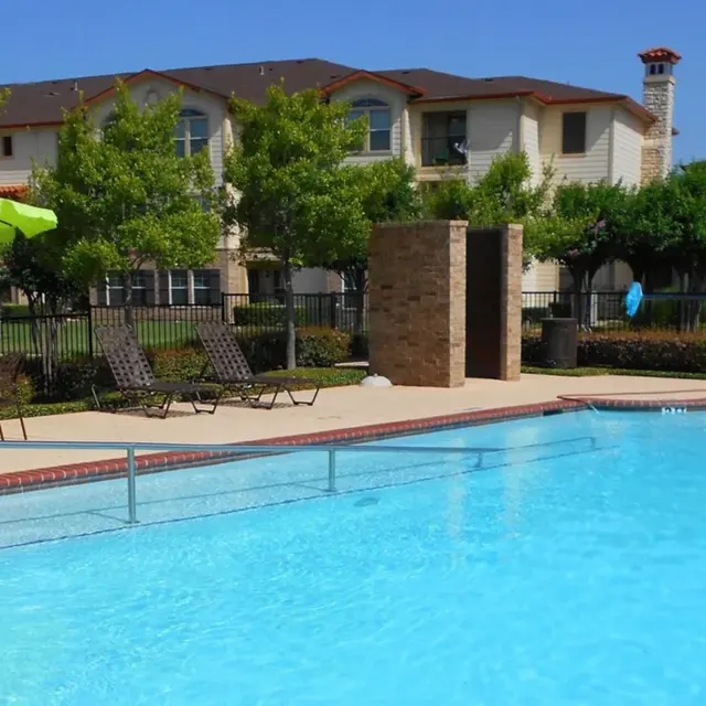 A swimming pool surrounded by lounge chairs and green trees, with an apartment building in the background under clear blue skies.