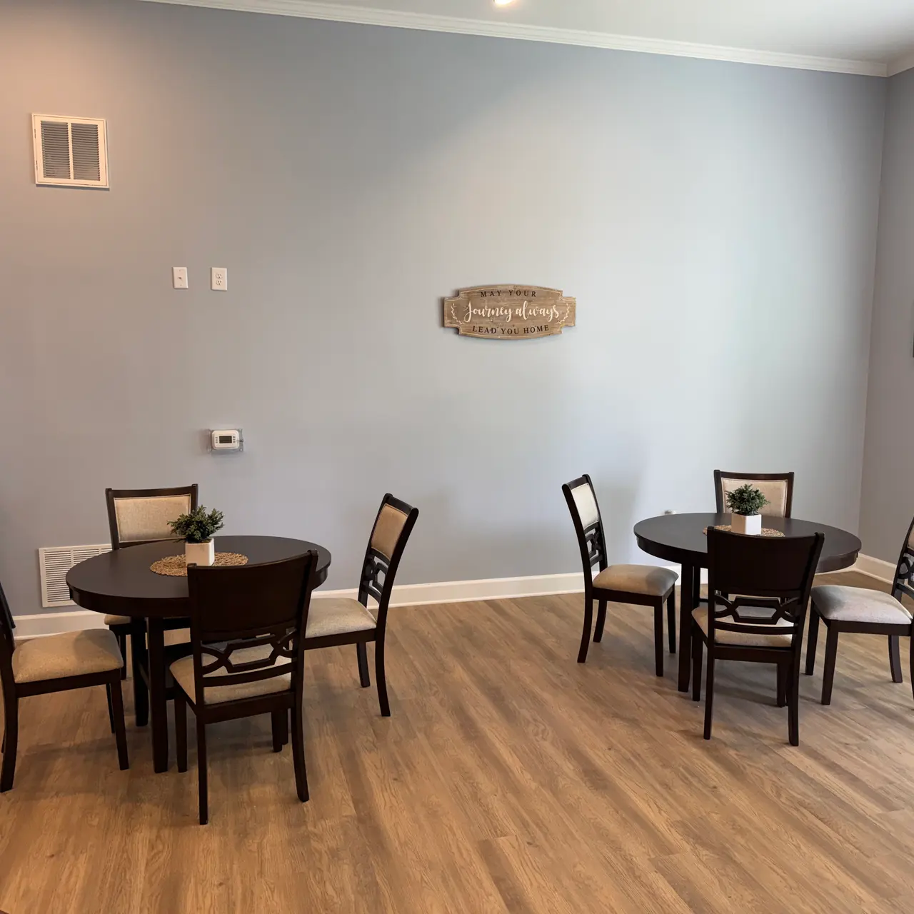 A spacious dining area featuring several round tables with chairs, adorned with small centerpieces. The walls are painted light blue, and there is a decorative wooden sign on the wall. The floor is made of light-colored wood.