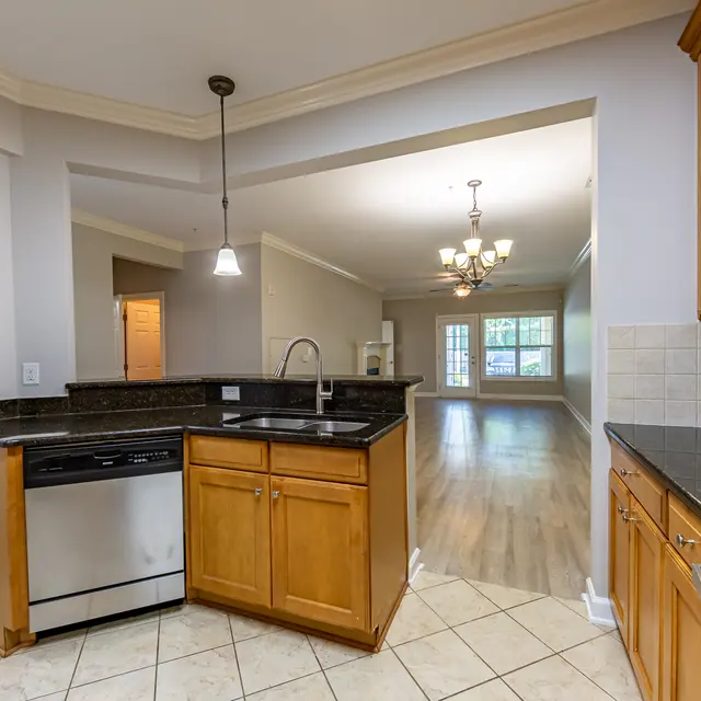 View of a modern kitchen with wooden cabinets and granite countertops, featuring stainless steel appliances and a view into a spacious living area.