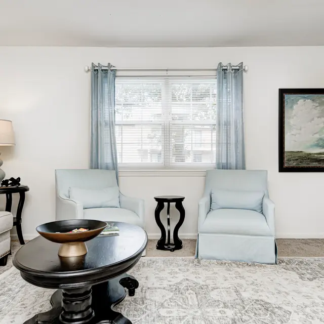 A cozy living room featuring two light blue chairs, a grey couch, and a dark round coffee table. There are light curtains on the windows and a painting on the wall. The space is bright and inviting.