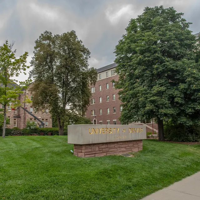 A view of a university campus featuring a grassy area, trees, and buildings under a cloudy sky.
