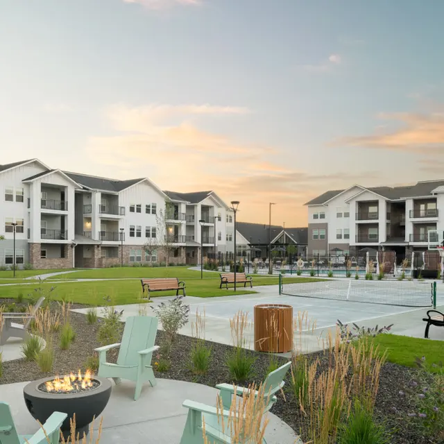 A modern residential complex with multiple buildings, featuring a landscaped outdoor area with benches, grass, and a fire pit.