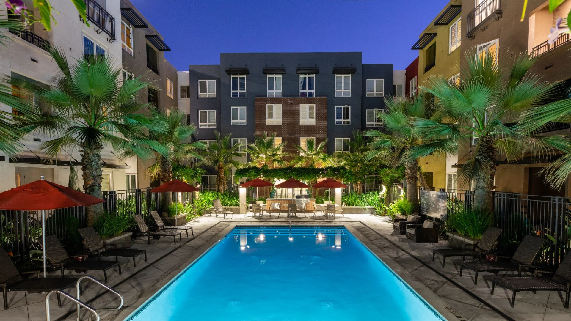 Evening Pool Scene in Apartment Community A well-lit swimming pool surrounded by palm trees and lounge chairs at night, showcasing a vibrant apartment community.