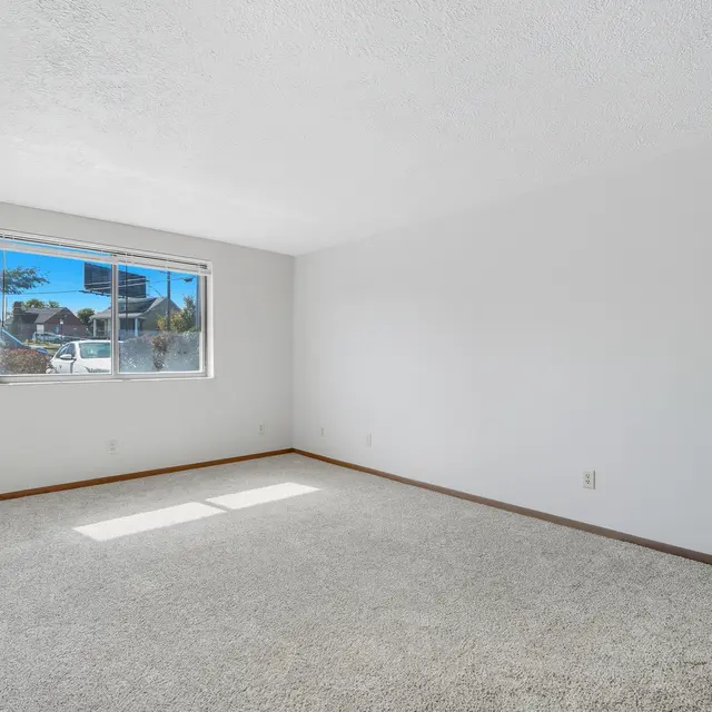 A spacious, empty room with light-colored walls and carpet. A window allows natural light in, with a view of a neighborhood outside. An air conditioning unit is visible to the left.
