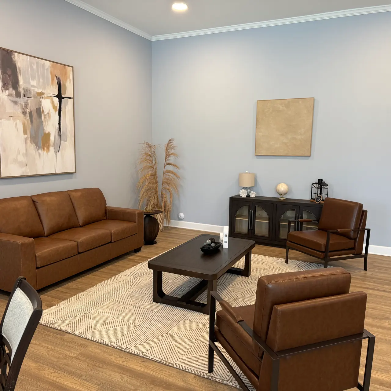 A modern living room featuring a brown leather sofa, two brown chairs, a coffee table, and a decorative area rug. There are light blue walls and abstract artwork hanging on the wall.