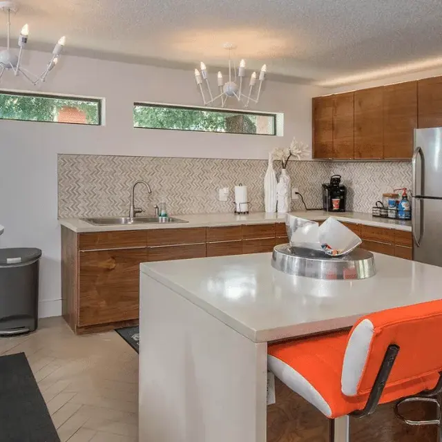 A modern kitchen featuring sleek wood cabinetry, gray countertops, stainless steel appliances, and stylish orange and white bar stools. The kitchen has a minimalist design with large windows allowing natural light to enter, and a contemporary chandelier above the island.