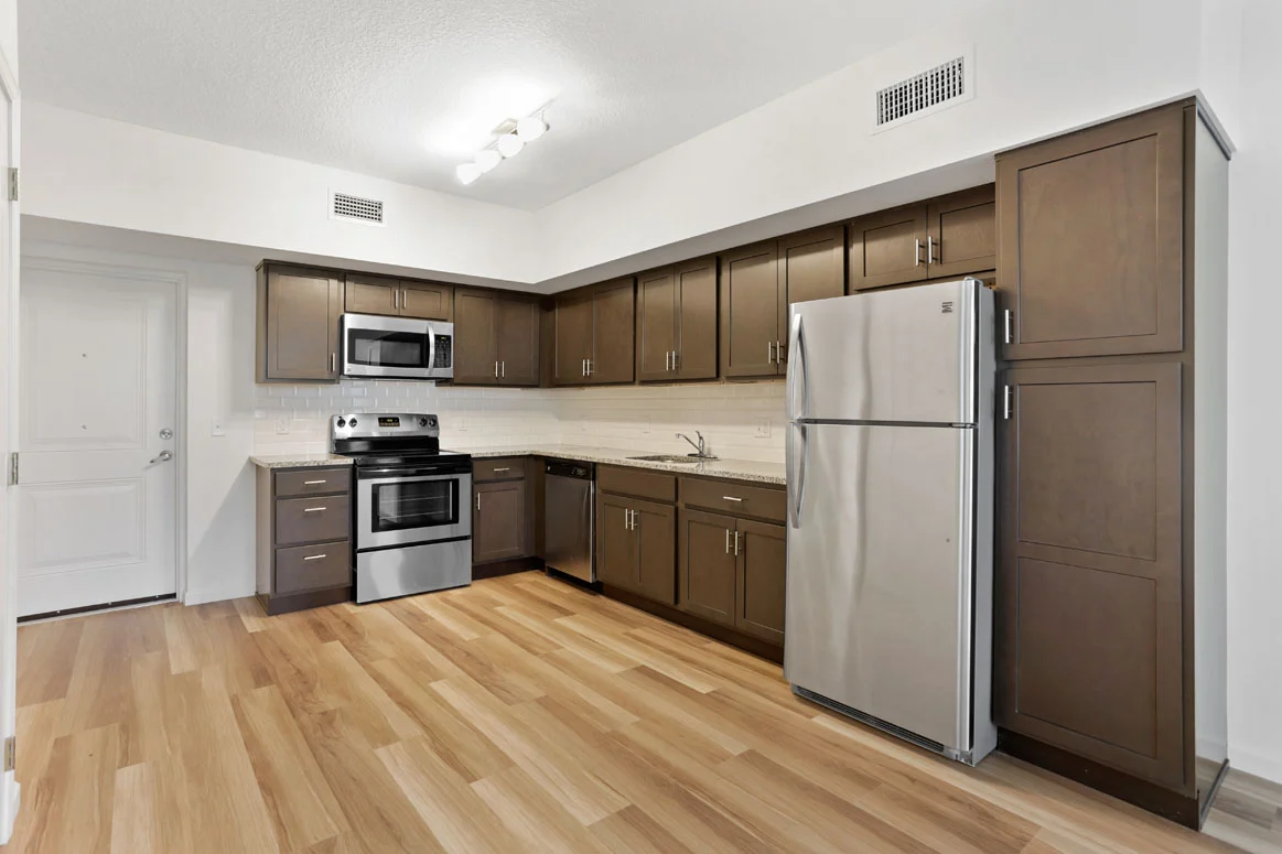 A modern kitchen with dark wood cabinets, stainless steel appliances including a stove and a refrigerator, and light wood flooring.