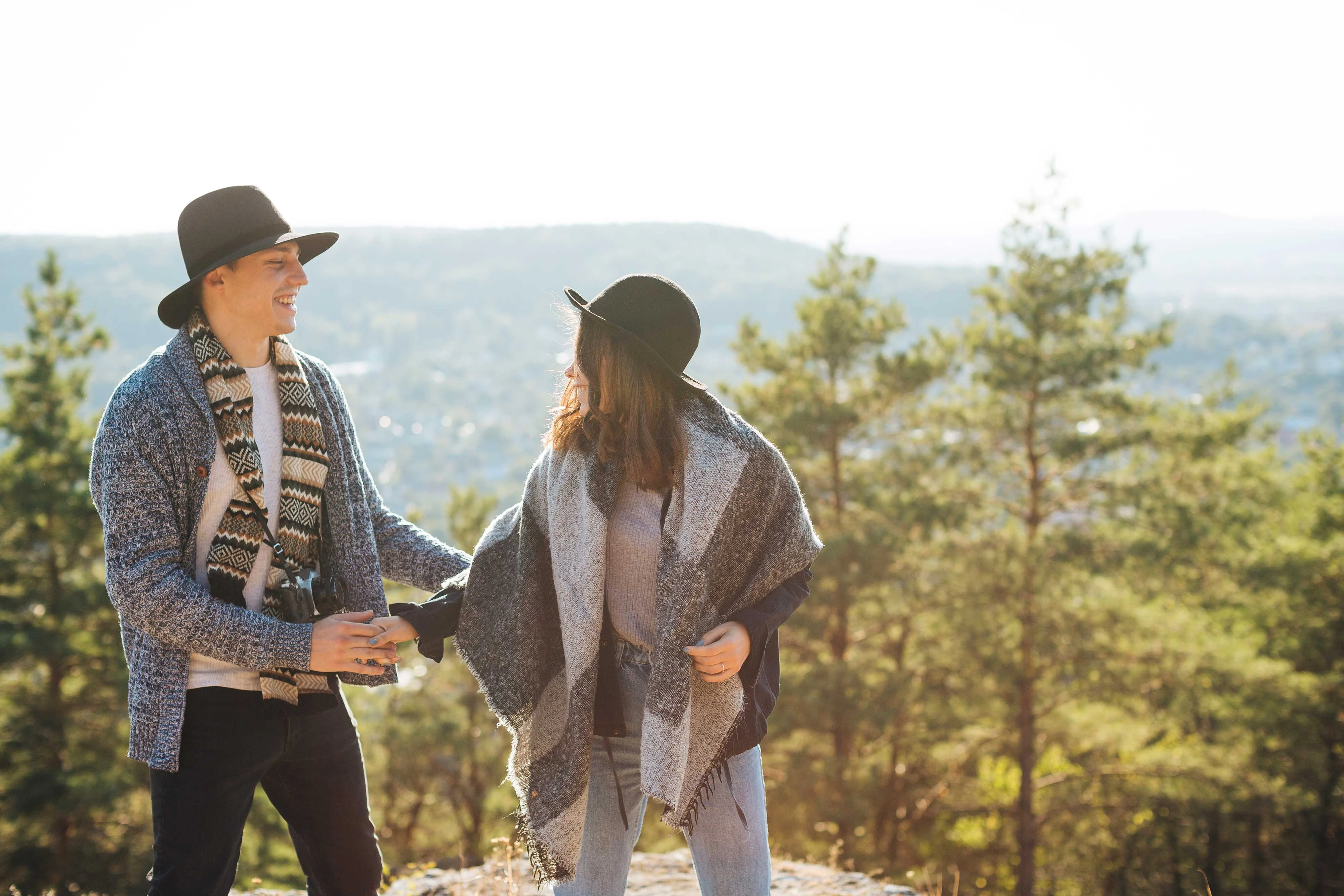A couple laughing and enjoying their time together in the mountains. One partner is wearing a blue sweater and hat, while the other is dressed in a grey poncho and black hat. They are engaged in a joyful moment, surrounded by trees and a scenic view in the background.