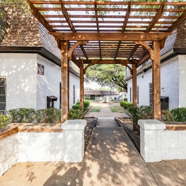 A walkway leading through a landscaped courtyard with white buildings on either side and a wooden pergola overhead.