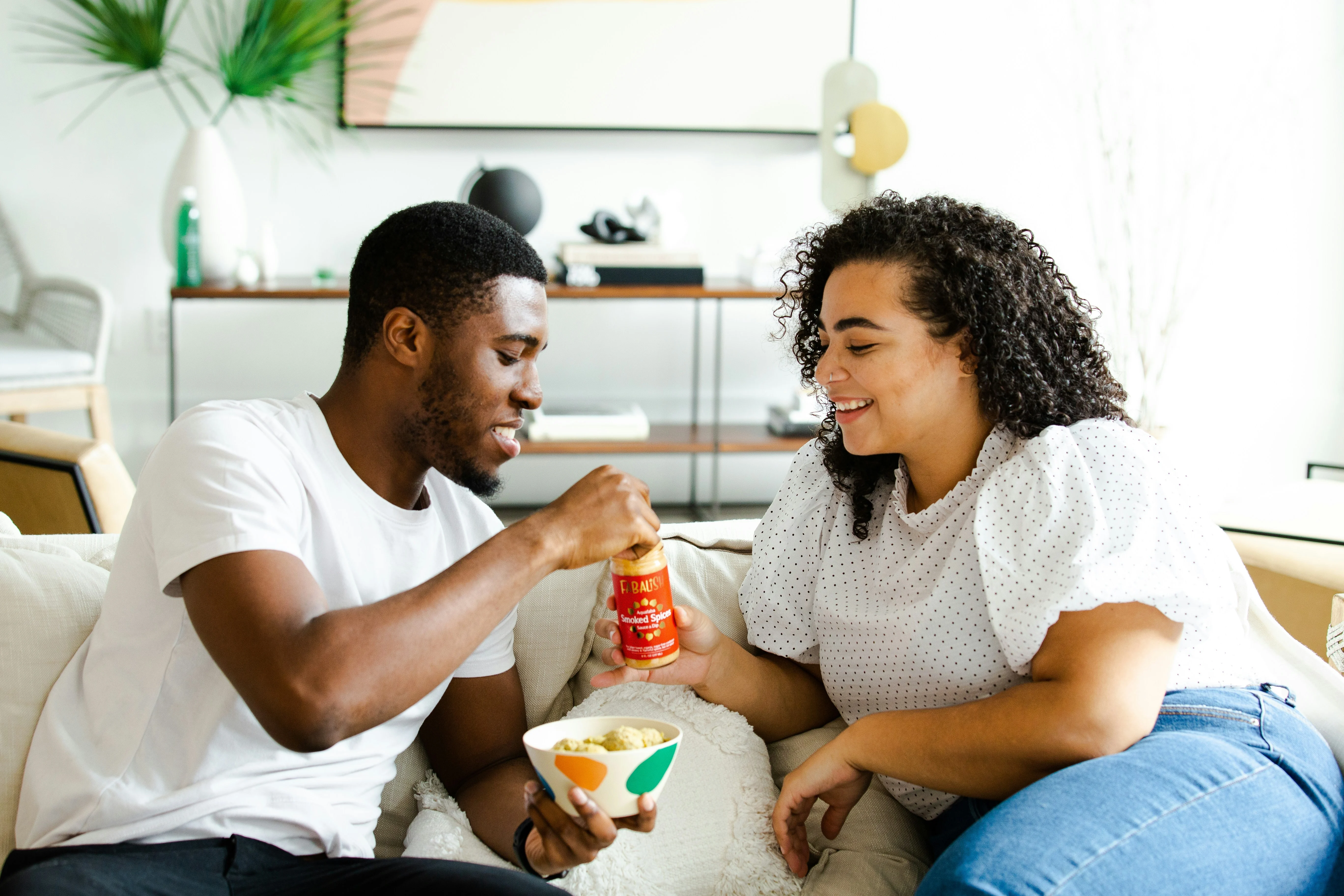 A couple sitting on a couch. The man is offering a jar of sauce to the woman, who is smiling. She has a bowl of food in her lap. The setting is bright and modern with plants in the background.