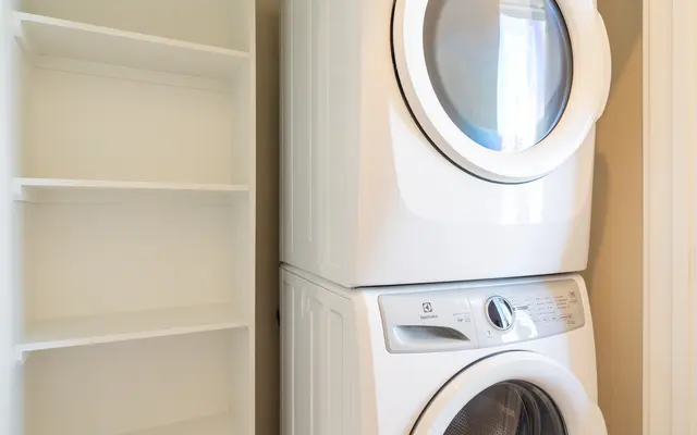 A compact laundry room featuring a stacked washer and dryer unit in white. Next to it is a tall, empty white shelving unit.