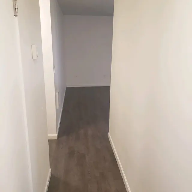 A narrow hallway with light-colored walls and dark wooden flooring, leading toward a bright area in the distance.