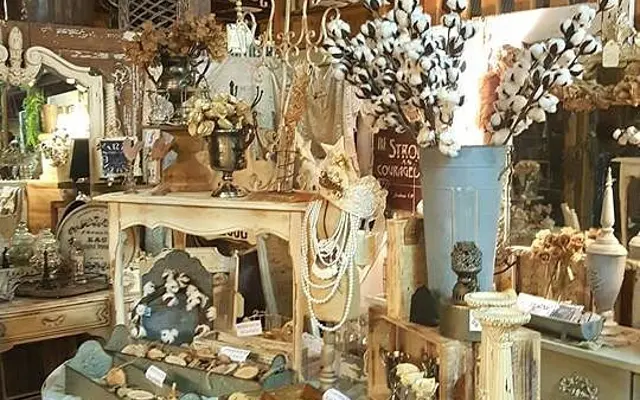 A charming interior filled with vintage home decor, showcasing a beautifully arranged table covered with various decorative items, a large chandelier overhead, and rustic signs on wooden walls.