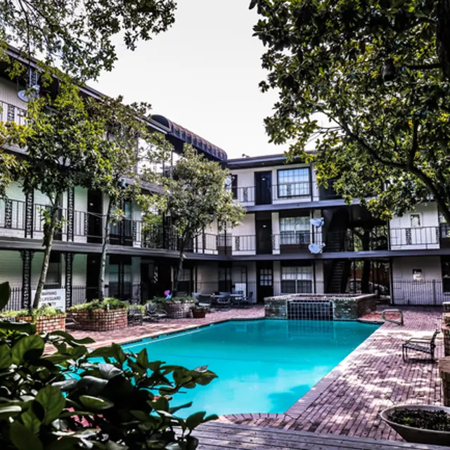 A courtyard view of an apartment complex featuring a blue swimming pool surrounded by greenery and brick pathways.