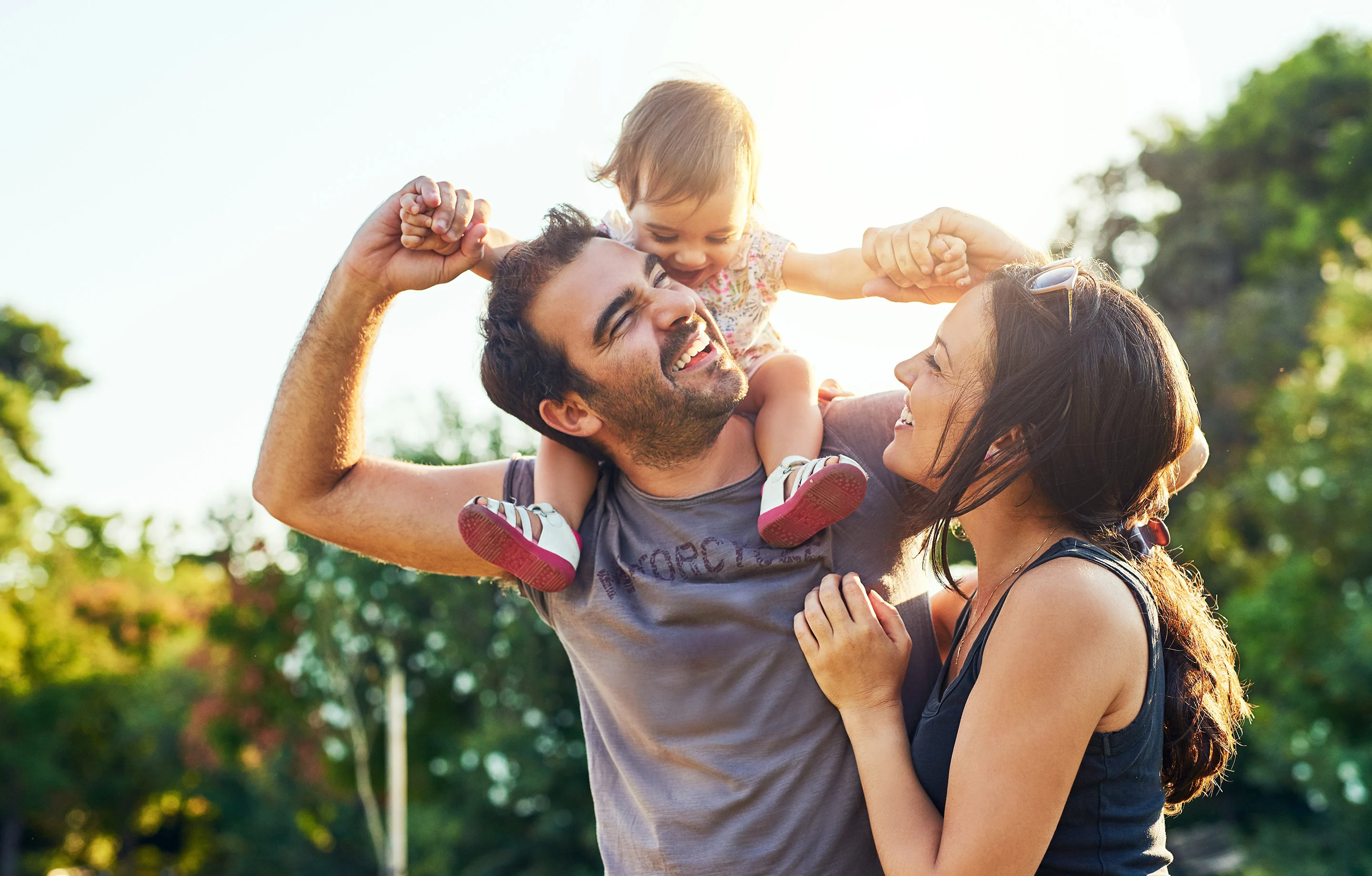 A joyful family playing outside in a sunny park. A father holds a small child on his shoulders, while the mother smiles beside them. The scene is lush with greenery.