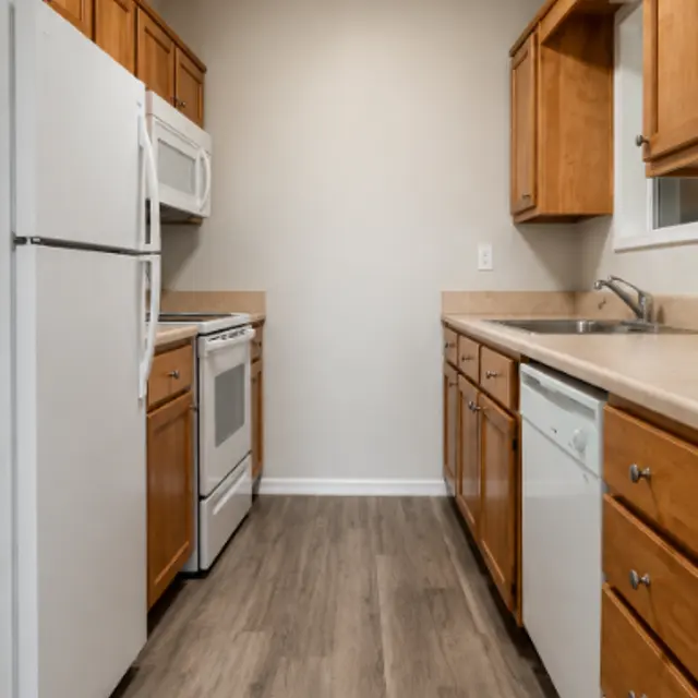 A narrow kitchen featuring wooden cabinets, stainless steel appliances including a refrigerator, microwave, and oven, with a light color countertop and a sink.