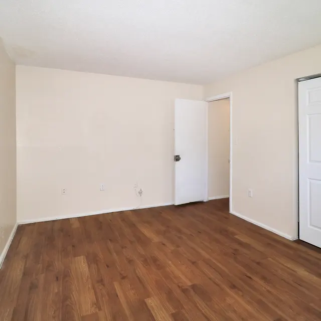 An empty room with light-colored walls and wooden flooring. There's a closed sliding wardrobe with white doors on the right and an open door leading to another room on the left.